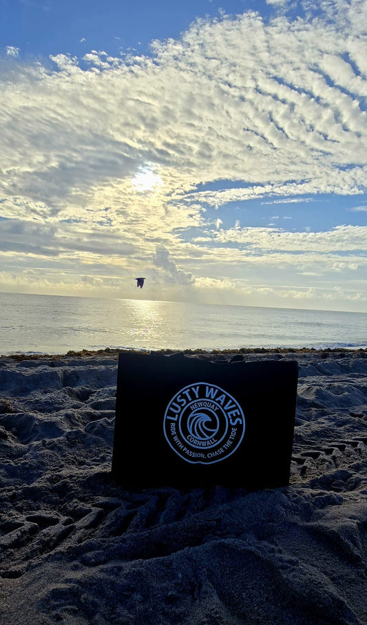 Black bag with 'Lusty Waves' logo on a sandy beach with ocean and cloudy sky in the background