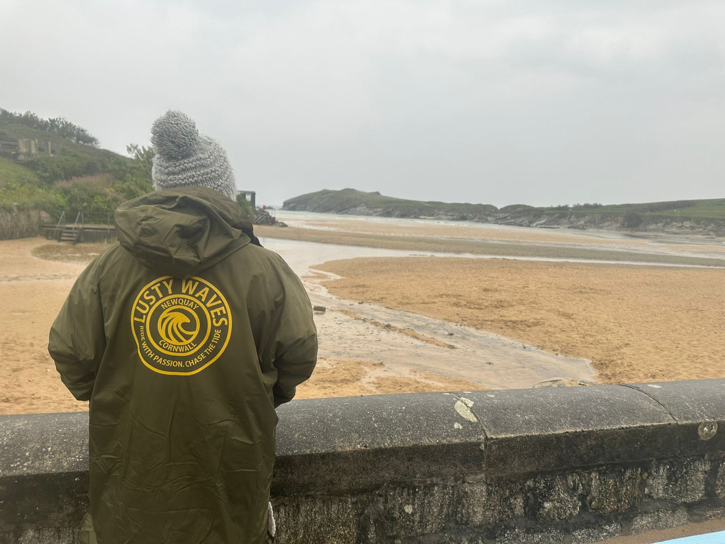 Person wearing a green jacket with a logo, standing on a bridge overlooking a beach.