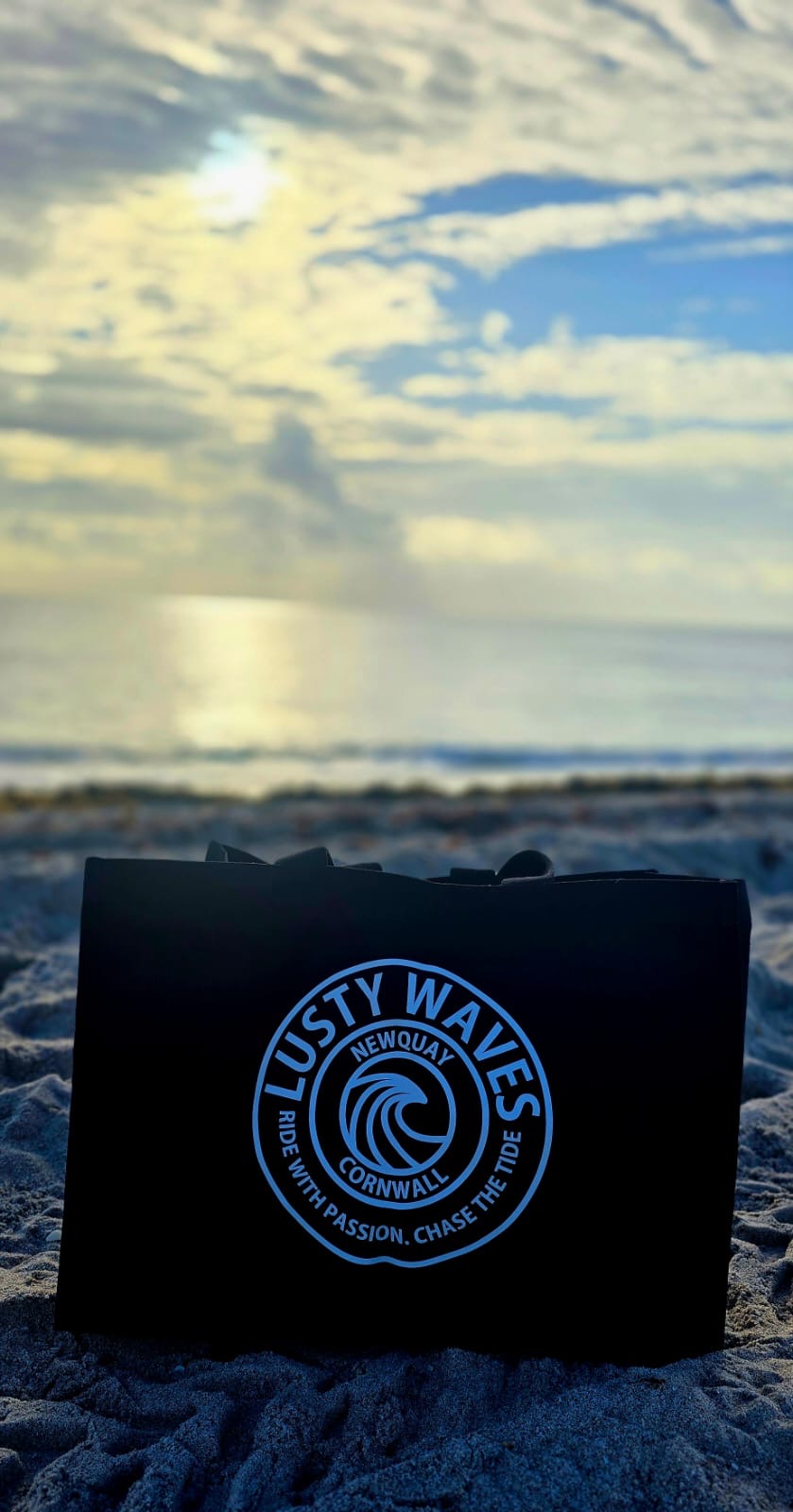 Black bag with 'Lusty Waves' logo on a beach with ocean and sky in the background